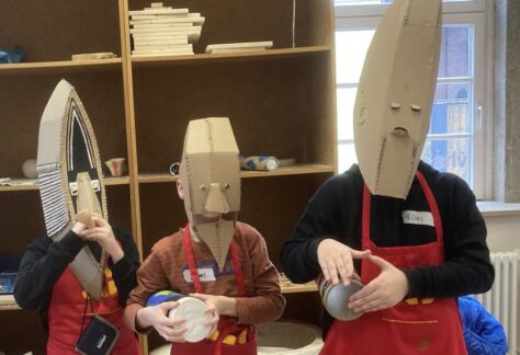 Stehende Kinder in roten Schürzen und mit gestalteten Masken aus Pappe spielen auf selbstgebauten Trommeln.