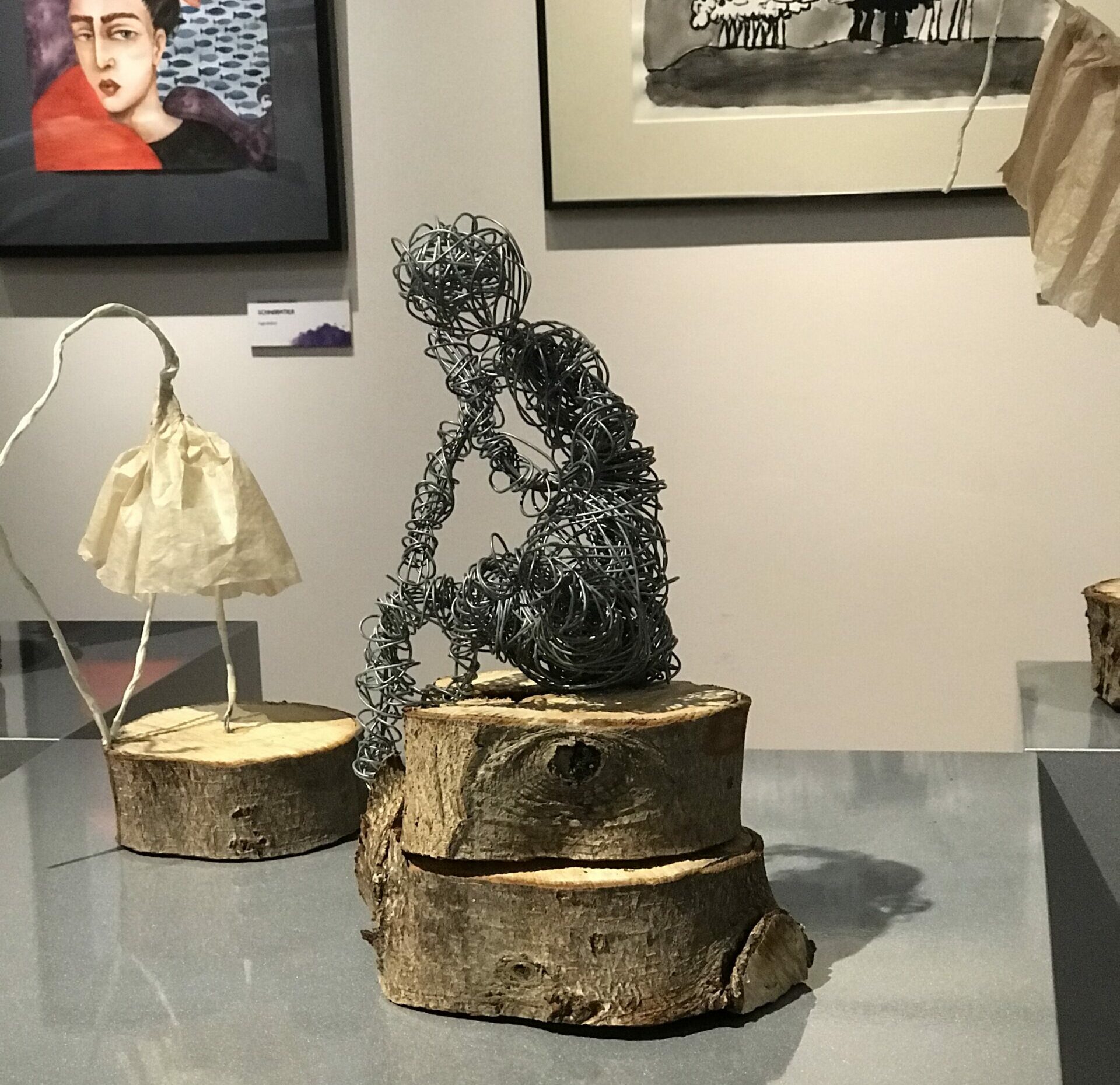Blick in eine Ausstellung, im Vordergrund aus Draht geformte, sitzende Plastik auf einem Sockel aus Holzscheiben.